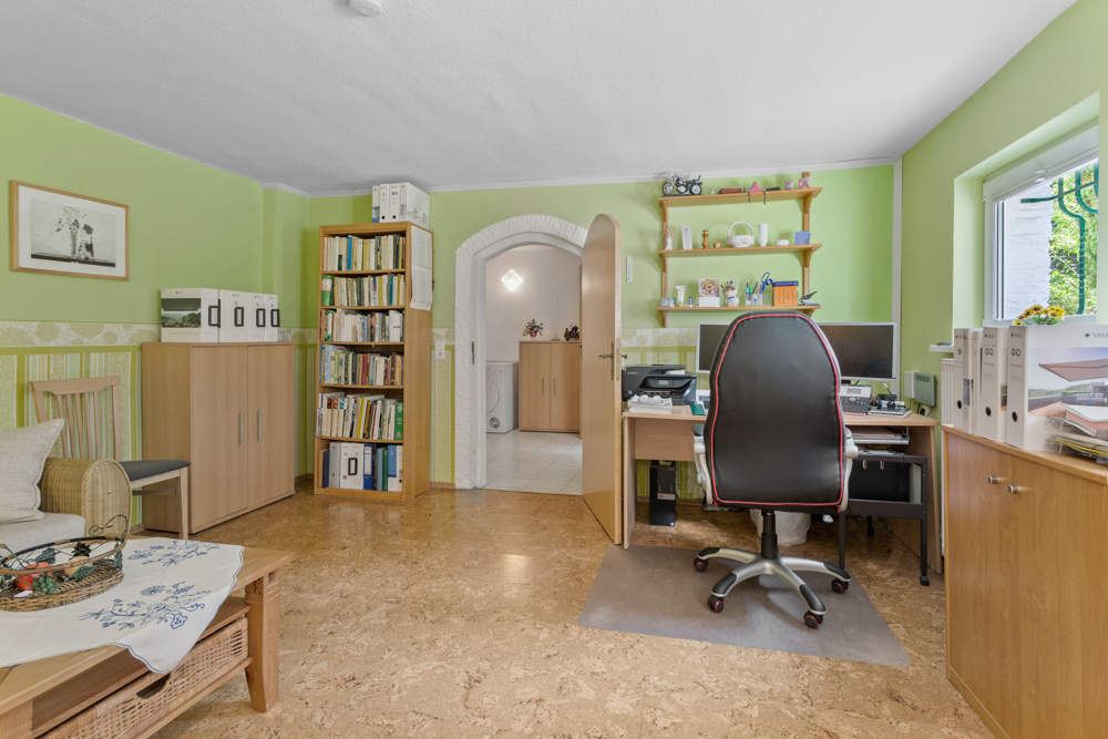 Helles Arbeitszimmer mit Schreibtisch, Bürostuhl, Regalen und Fenster, grüne Wände, Holzboden.