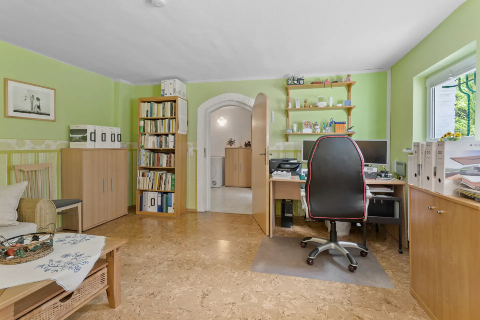 Helles Arbeitszimmer mit Schreibtisch, Bürostuhl, Regalen und Fenster, grüne Wände, Holzboden.