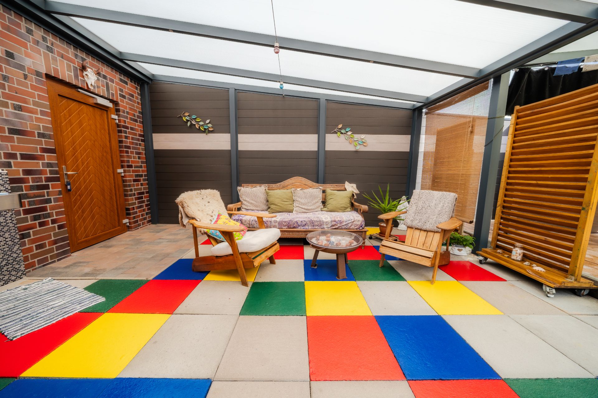 Überdachte Terrasse mit buntem Boden, Holzmöbeln, Sofa und Pflanzen.