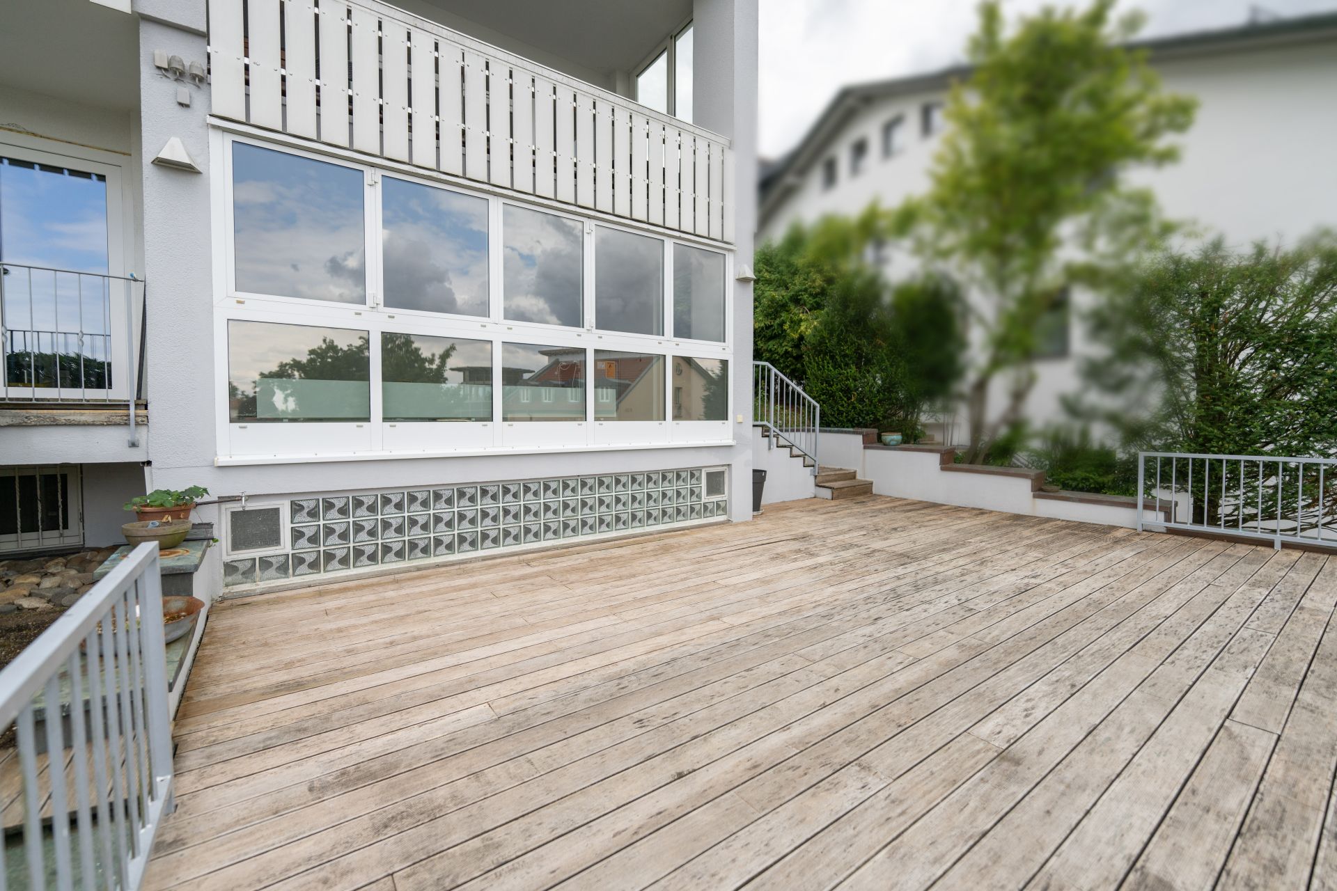 Große Holzterrasse mit Geländer an einem Wohnhaus, bepflanzte Beete und Treppe im Hintergrund.