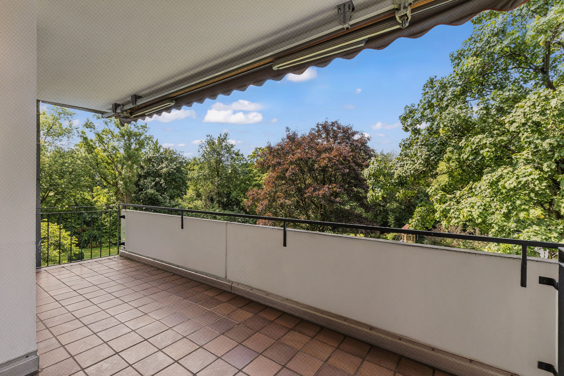 Überdachter Balkon mit braunen Fliesen und Blick auf grüne Bäume.
