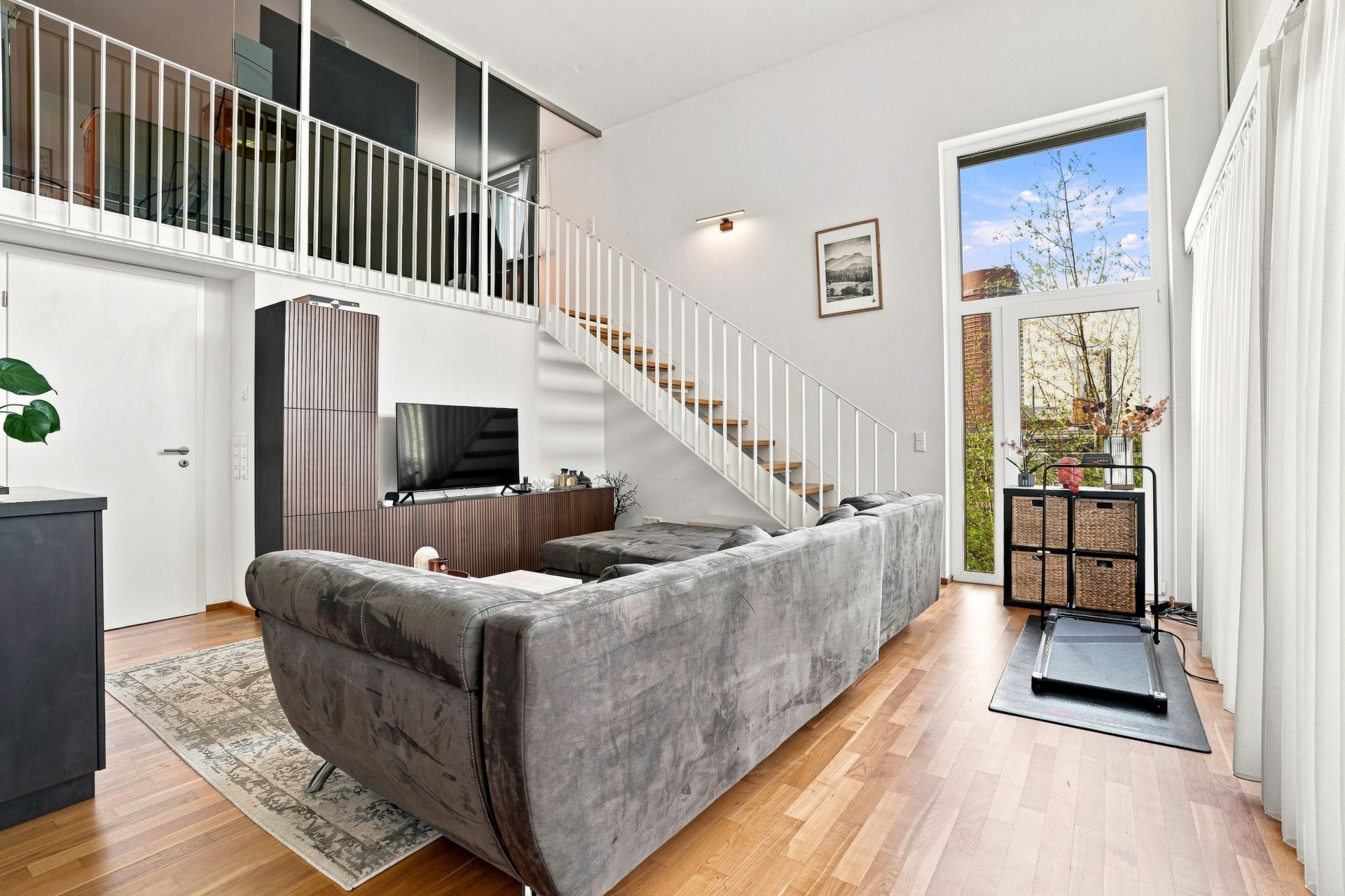 Wohnzimmer mit grauem Sofa, Fernseher, Treppe zur Galerie und großen Fenstern mit Blick nach draußen.