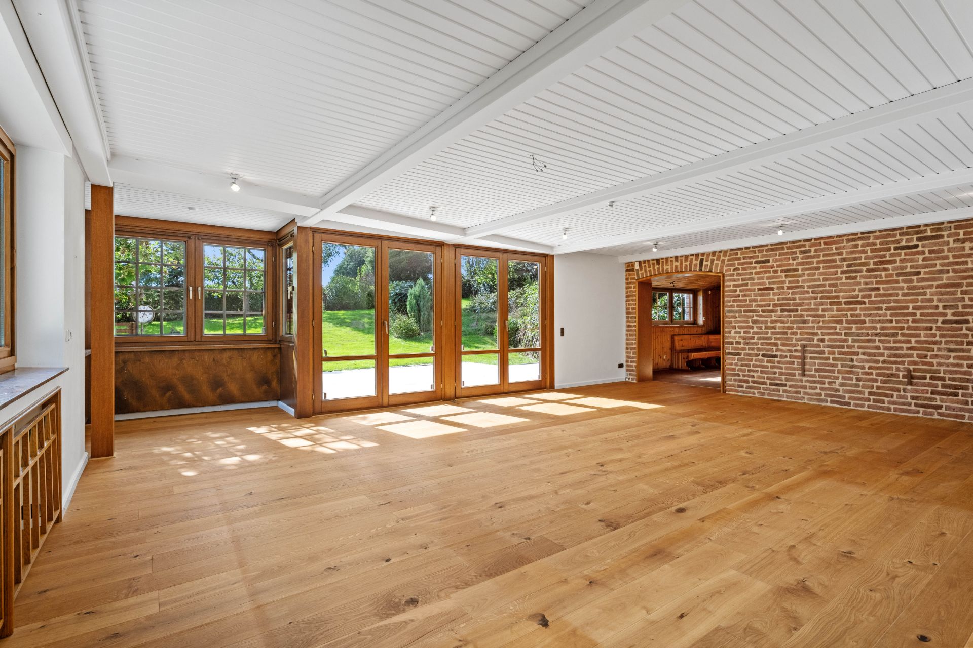 Heller Raum mit Holzboden, großen Fenstern und einer Backsteinwand, Blick auf den Garten.