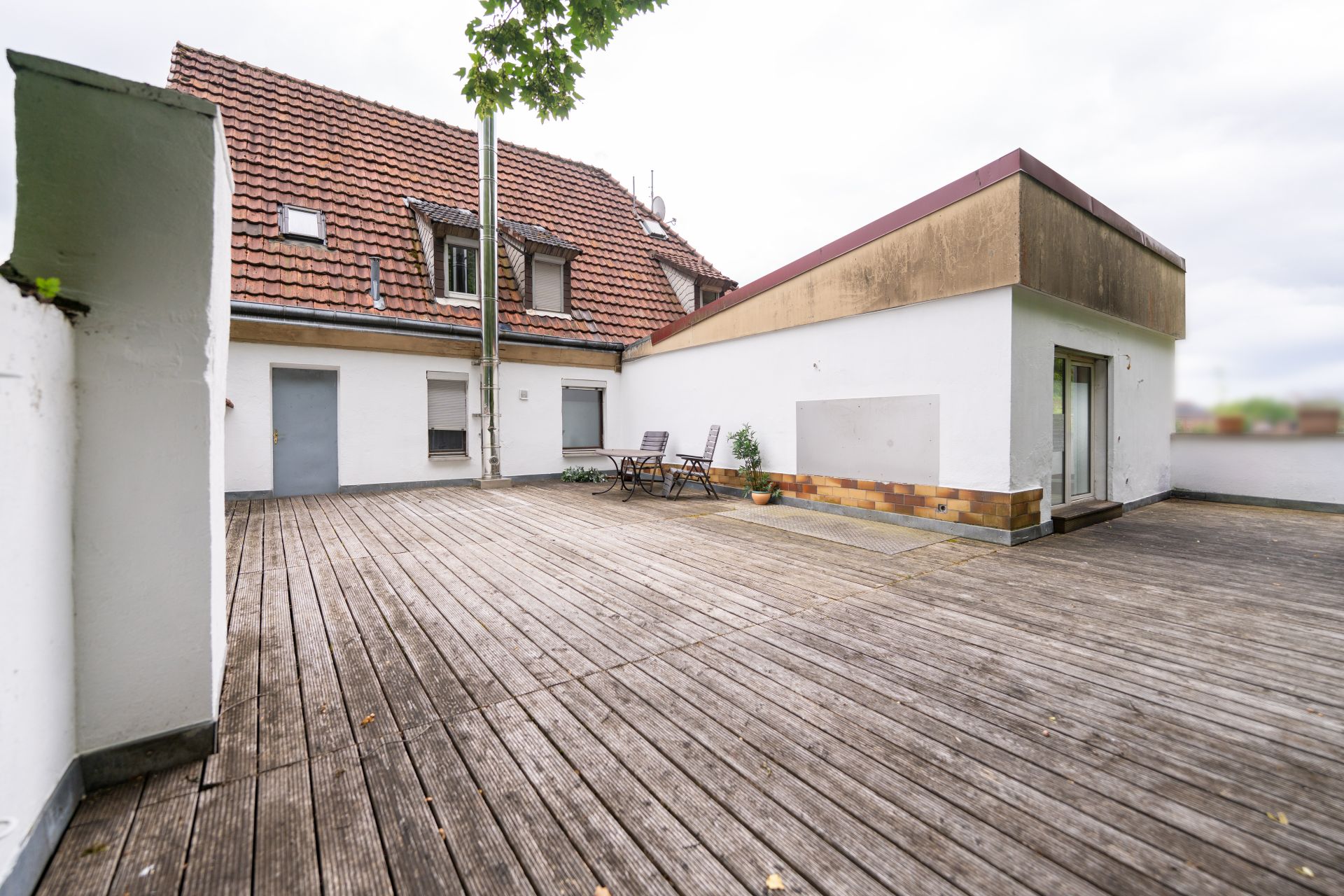 Große Dachterrasse mit Holzboden, Gartenmöbeln und Blick auf ein Wohnhaus mit rotem Dach.