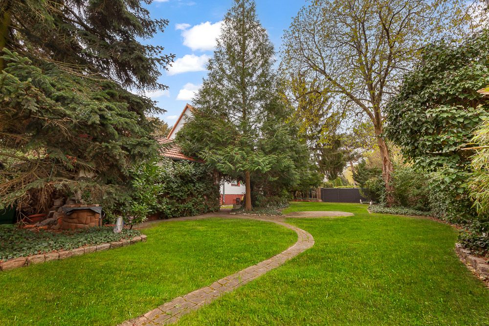Garten mit Rasenfläche, geschwungenem Weg und vielen Bäumen, Haus im Hintergrund.