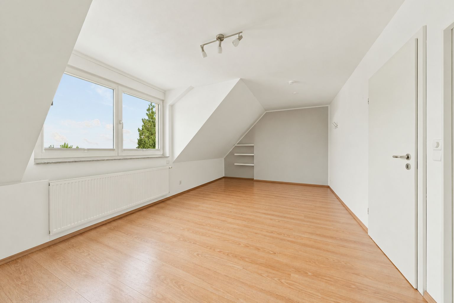 Helles Zimmer mit schräger Decke, großem Fenster, Holzfußboden und Regal in der Ecke.