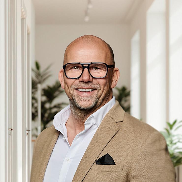 Ein lächelnder Mann mit Brille und beigem Sakko steht in einem hellen Flur mit Pflanzen.