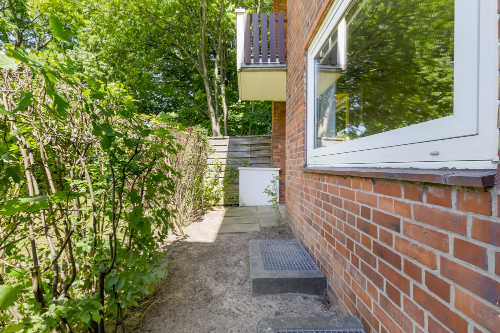 Schmaler Gartenweg mit Backsteinfassade, Hecke und großem Fenster.