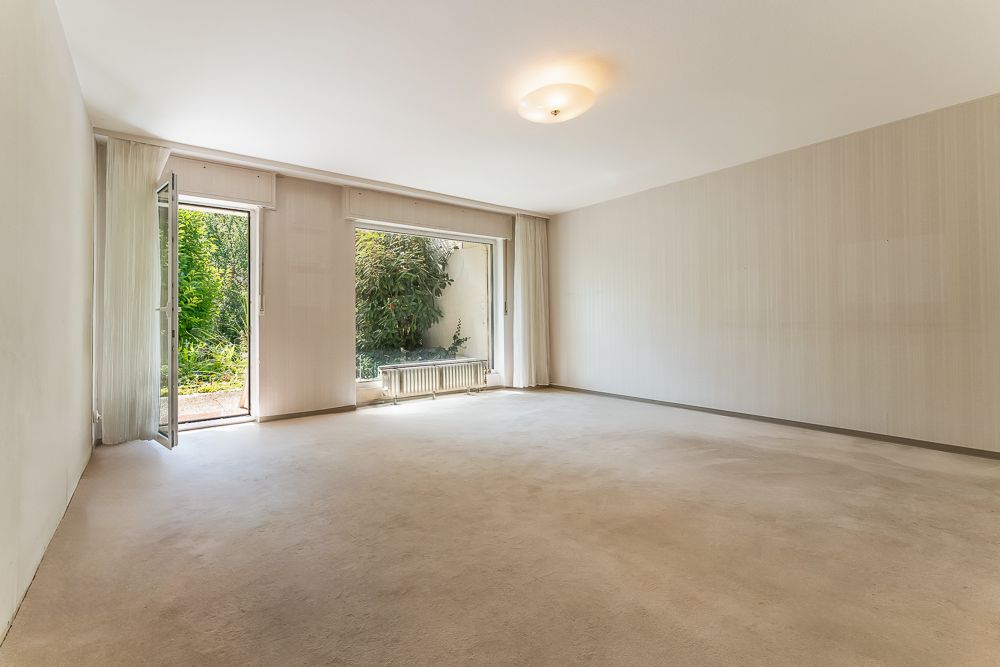 Leerer, heller Raum mit Teppichboden, großen Fenstern und offener Tür zum Garten.