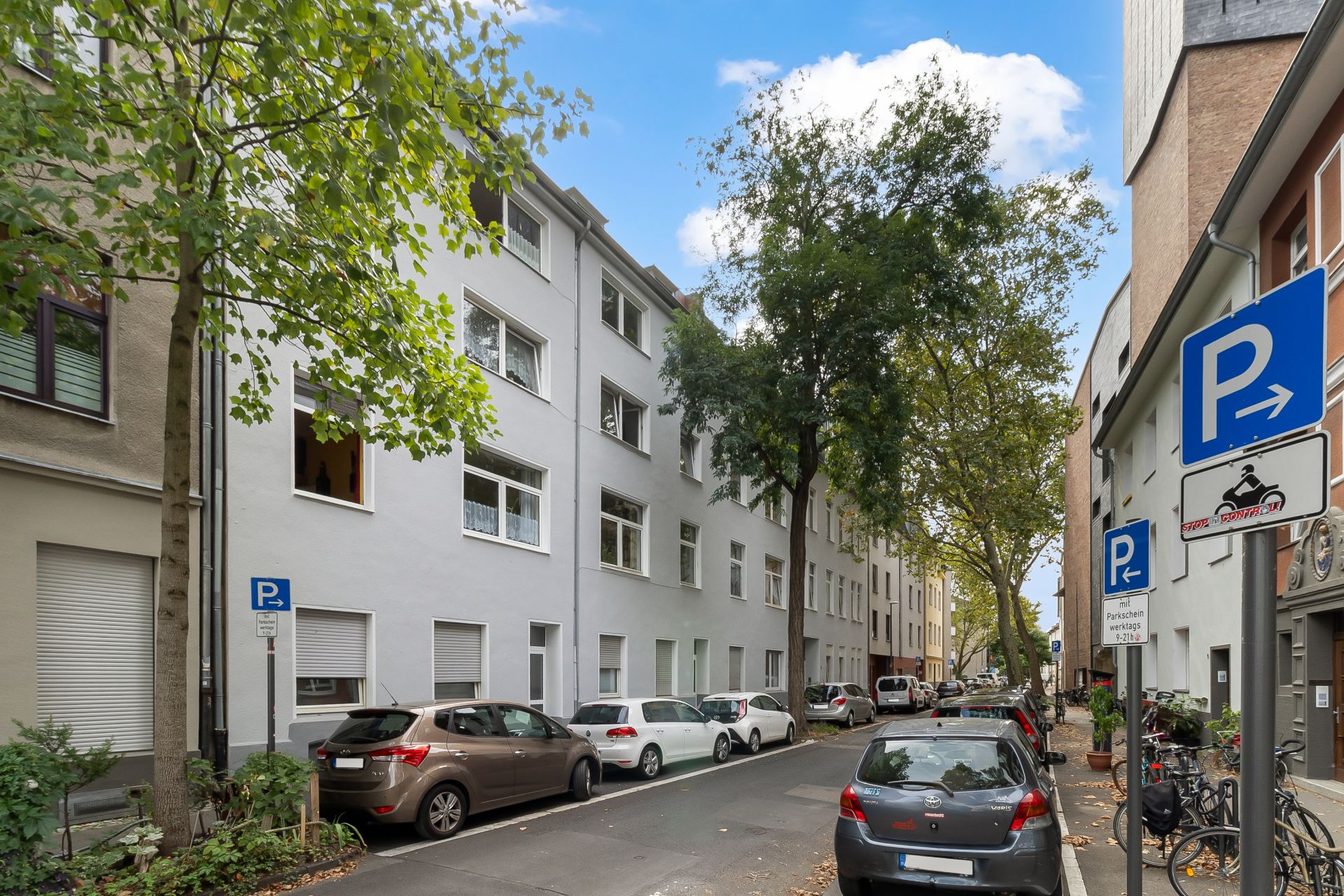 Wohnstraße mit geparkten Autos, Bäumen und mehrstöckigen Wohnhäusern bei Tag.