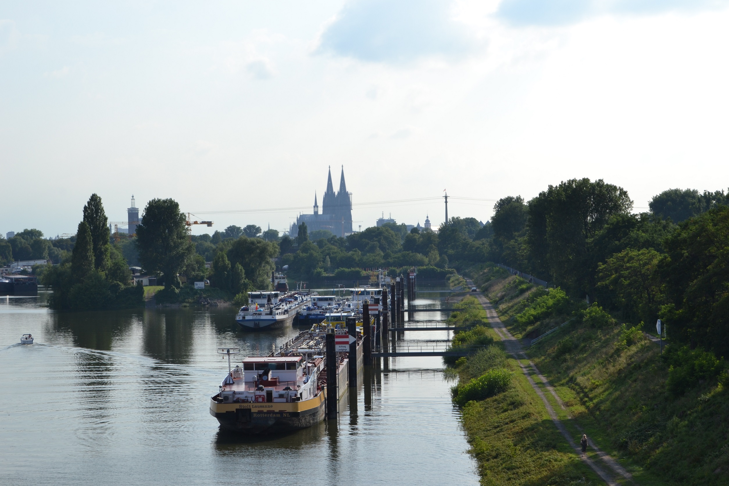 Frachtschiffe und Boote auf einem ruhigen Fluss mit Bäumen und Weg am Ufer.