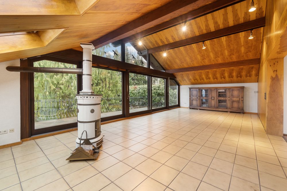 Großes, helles Wohnzimmer mit Fliesenboden, Holzdecke, Kaminofen und Fenstern zur Terrasse.