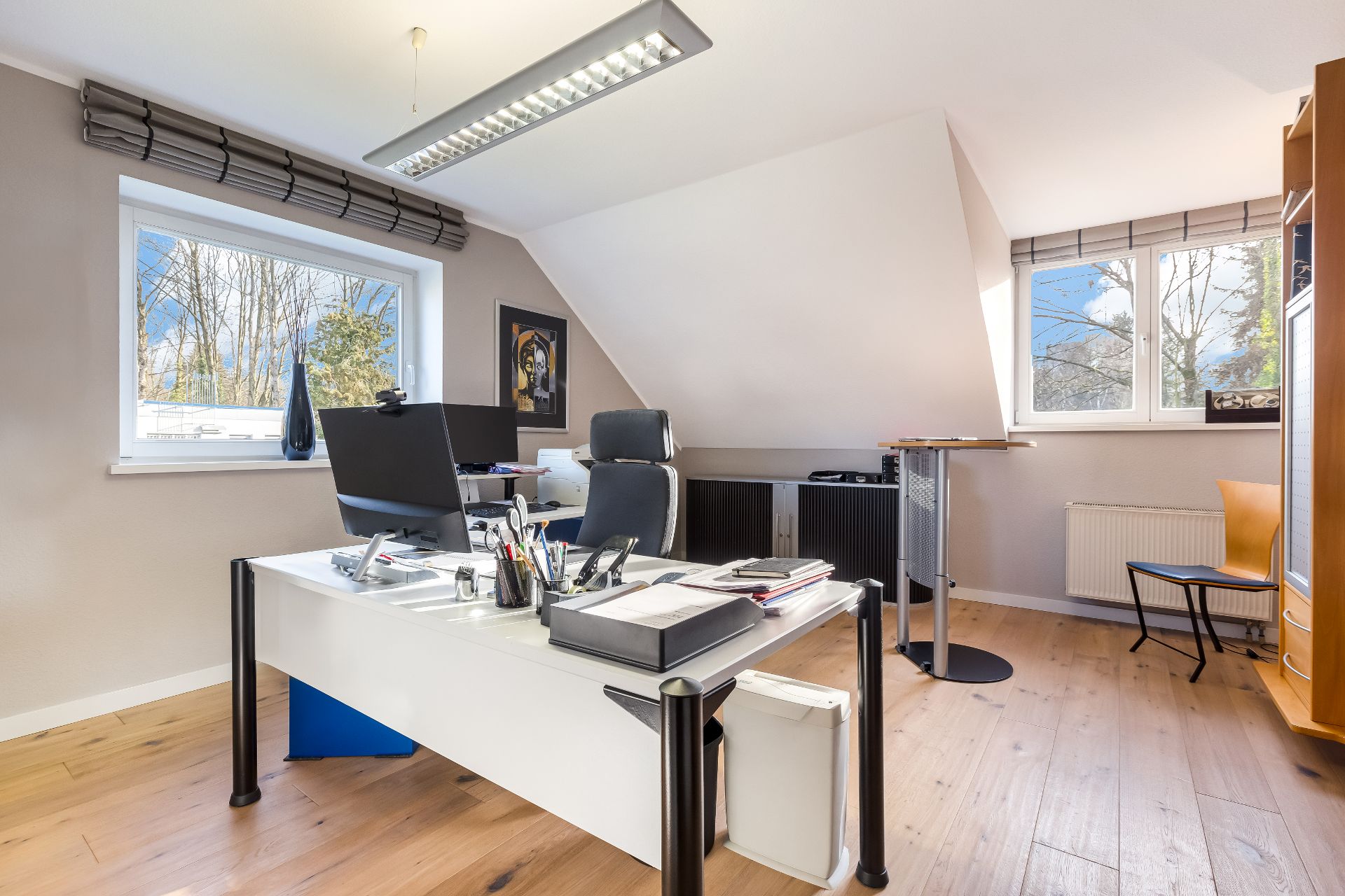 Helles Arbeitszimmer mit Schreibtisch, Monitoren, Stuhl und großen Fenstern auf Holzboden.