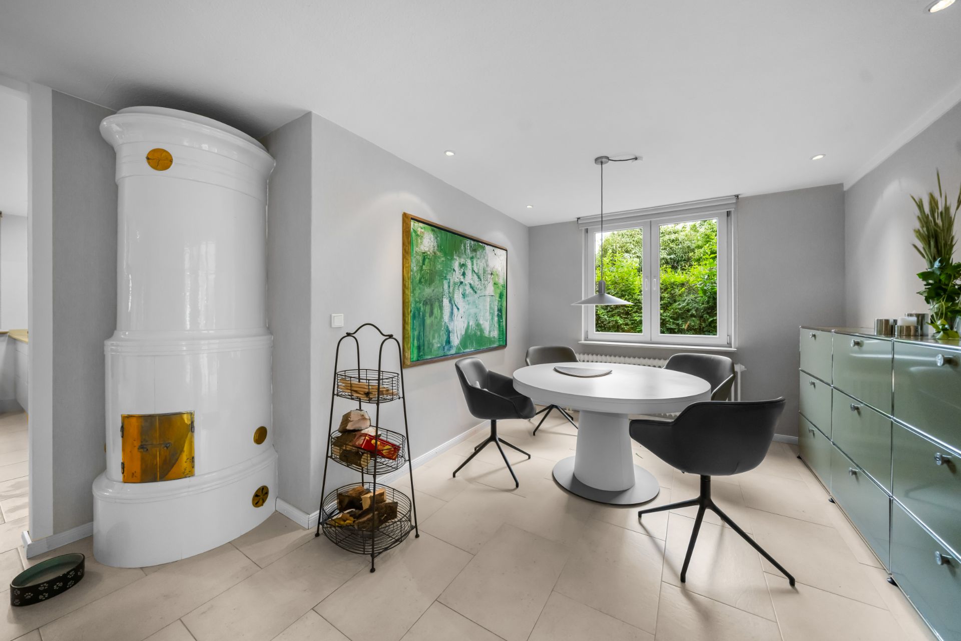 Modernes Esszimmer mit rundem Tisch, vier Stühlen, großem Bild, Ofen und Fenster mit Blick ins Grüne.