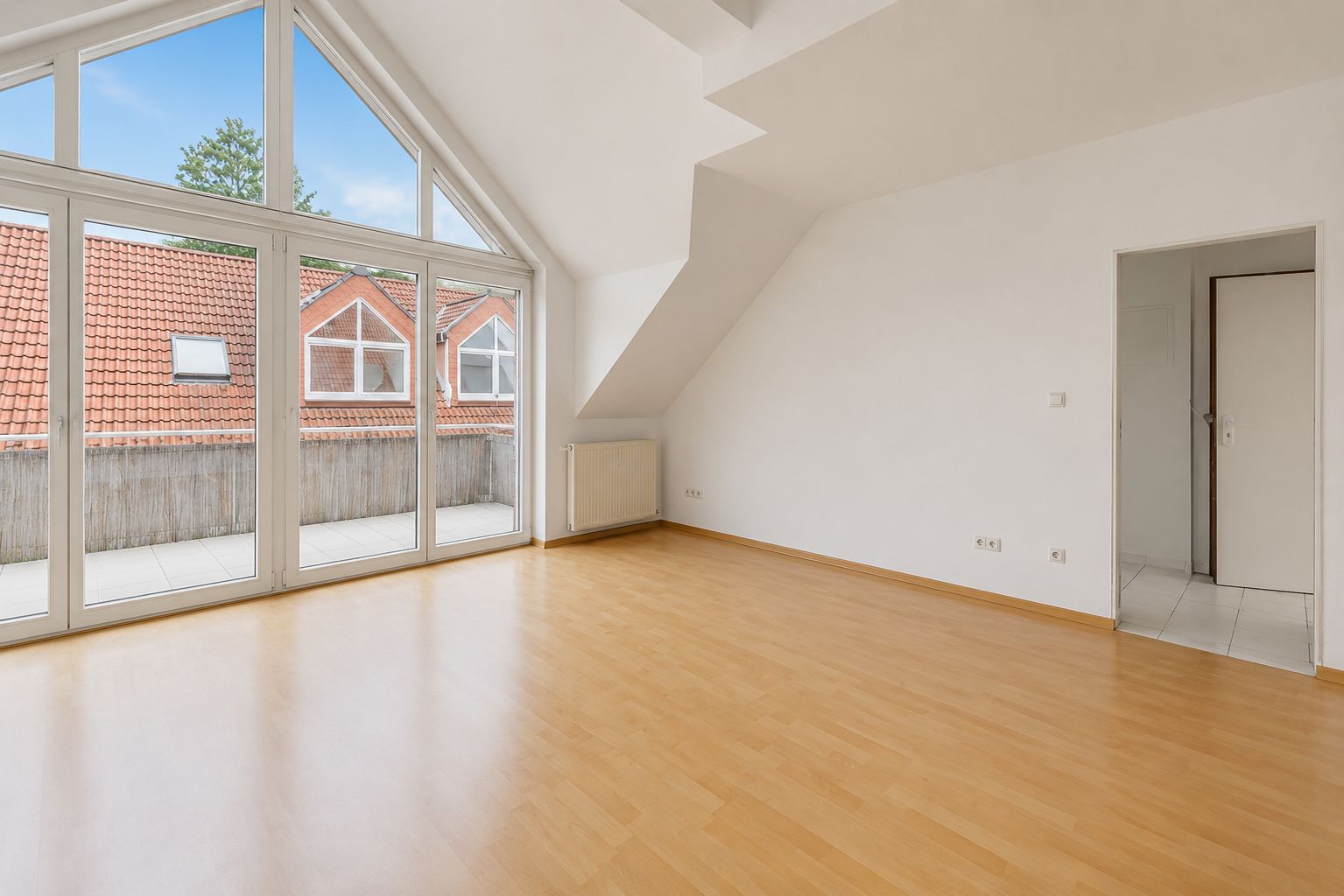 Leerer Raum mit Holzfußboden, großen Fenstern und Zugang zu einem Balkon.