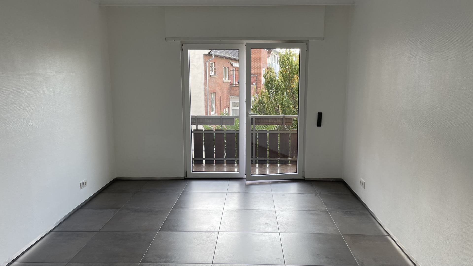 Leeres Zimmer mit dunklem Fliesenboden und Glastür zu einem kleinen Balkon.