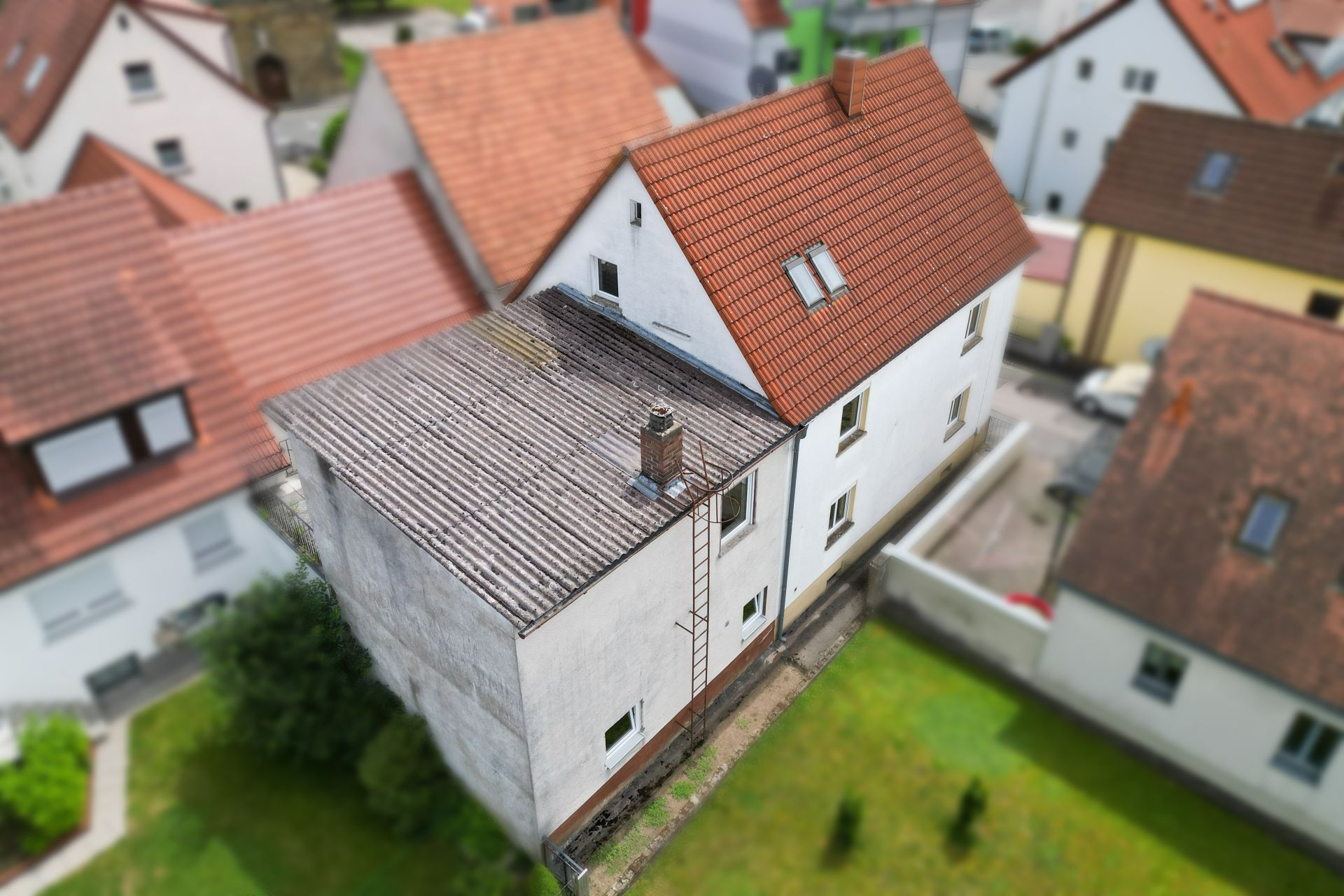 Weißes Wohnhaus mit rotem Schrägdach und Flachdachanbau, umgeben von weiteren Häusern.