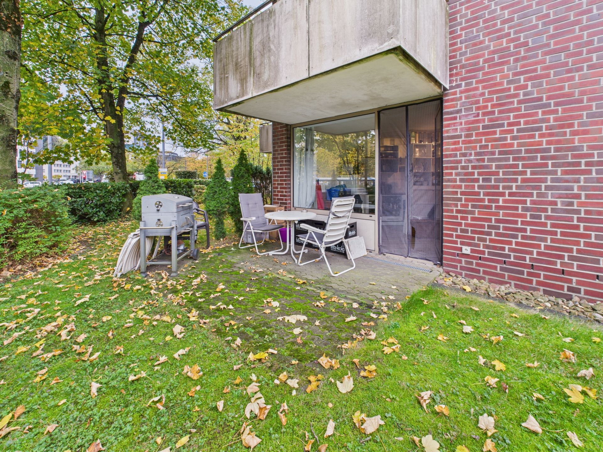 Terrasse mit Tisch, Stühlen und Grill an einer Wohnung mit Backsteinwand, umgeben von grünem Rasen und Bäumen.