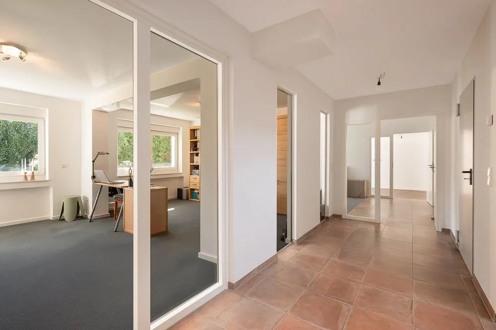 Heller Flur mit Fliesenboden und Glastüren, angrenzendes Zimmer mit Schreibtisch und Regalen.