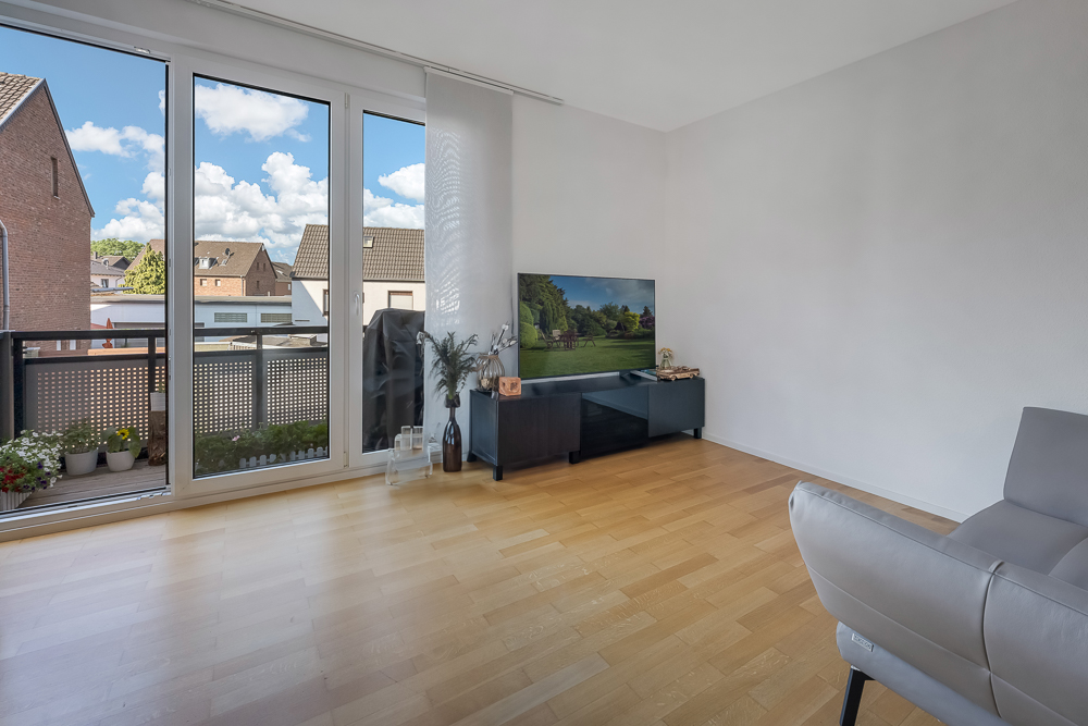 Helles Wohnzimmer mit Parkettboden, Sofa, TV auf Lowboard und Zugang zu einem Balkon mit Blumen.
