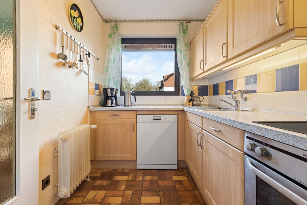 Helle Küche mit Holzschränken, Geschirrspüler, Herd und Fenster mit Blick nach draußen.