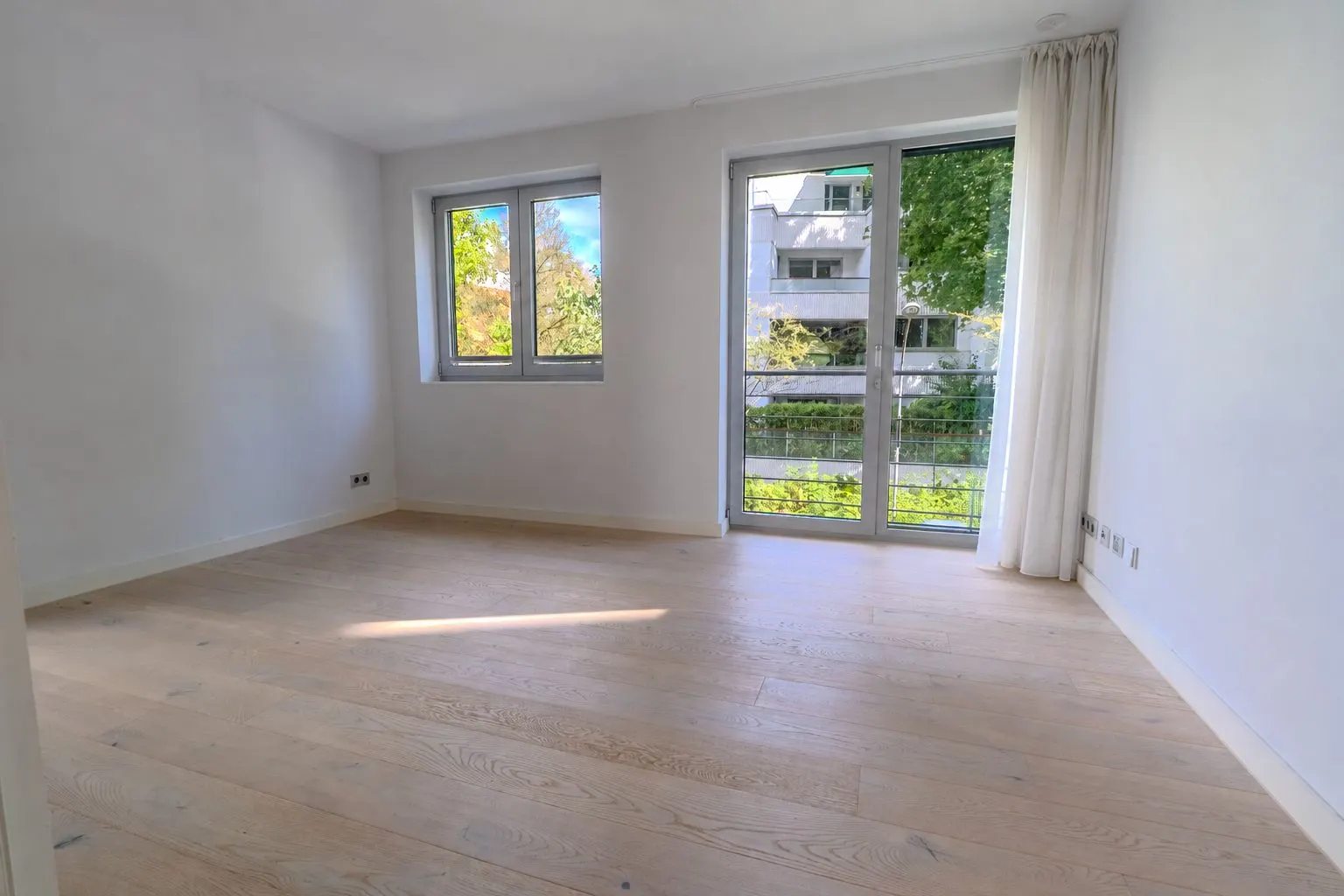 Helles, leeres Zimmer mit Holzfußboden, zwei Fenstern und einer Glastür mit Blick ins Grüne.