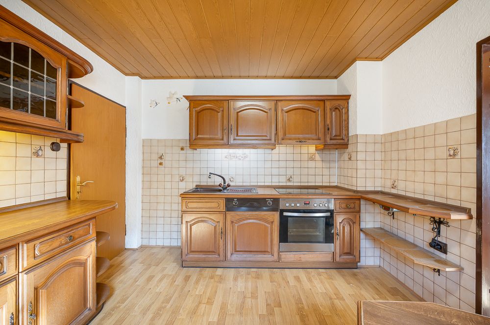 Küche mit Holzschränken, Elektroherd und Fliesenwand, Holzboden und Regalen an der rechten Wand.