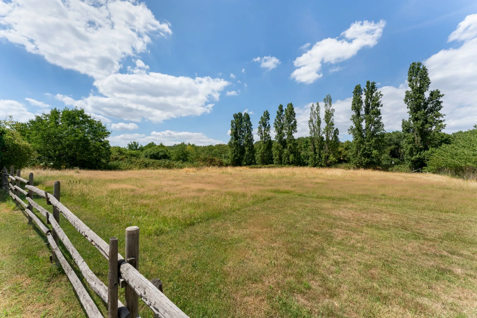 Große Wiese mit Holzzaun und Bäumen im Hintergrund unter blauem Himmel.