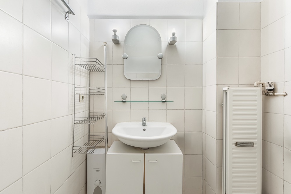 Kleines Badezimmer mit Waschbecken, Spiegel, Regal und Heizkörper, alles in weißen Fliesen.