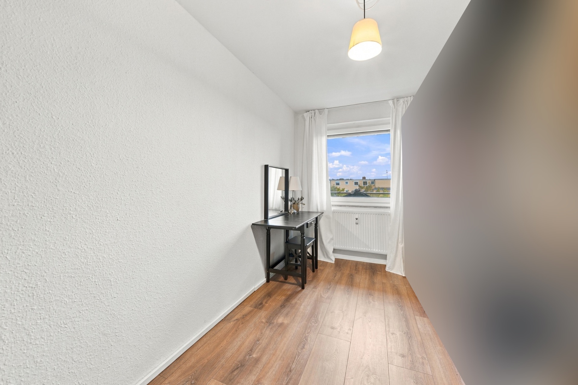 Schmaler Raum mit Holzboden, Schminktisch, Stuhl und Fenster mit Blick nach draußen.