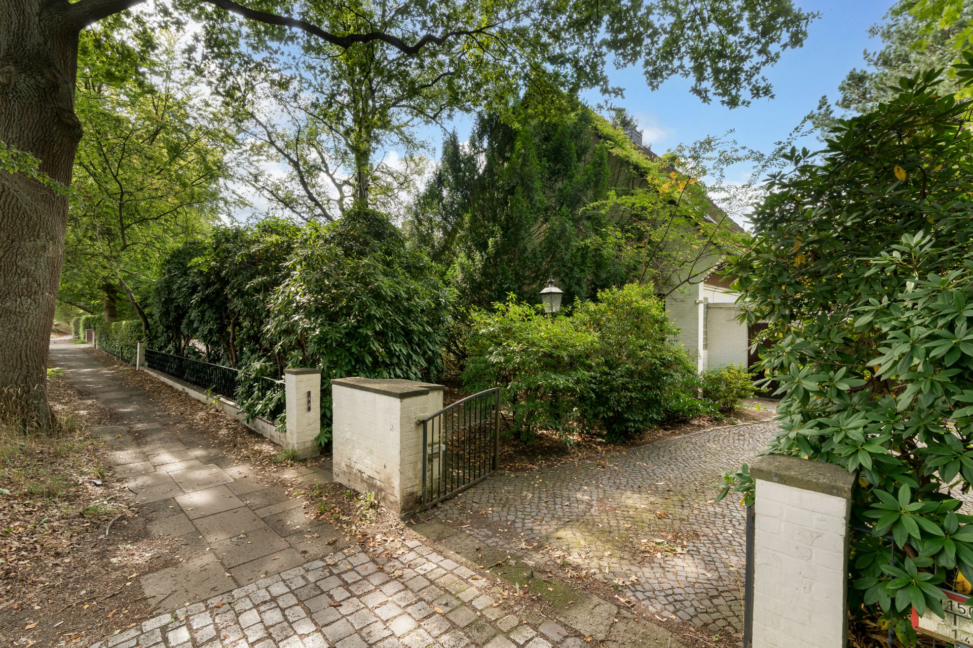 Eingangsbereich eines Hauses mit Tor, gepflastertem Weg und viel grüner Bepflanzung.