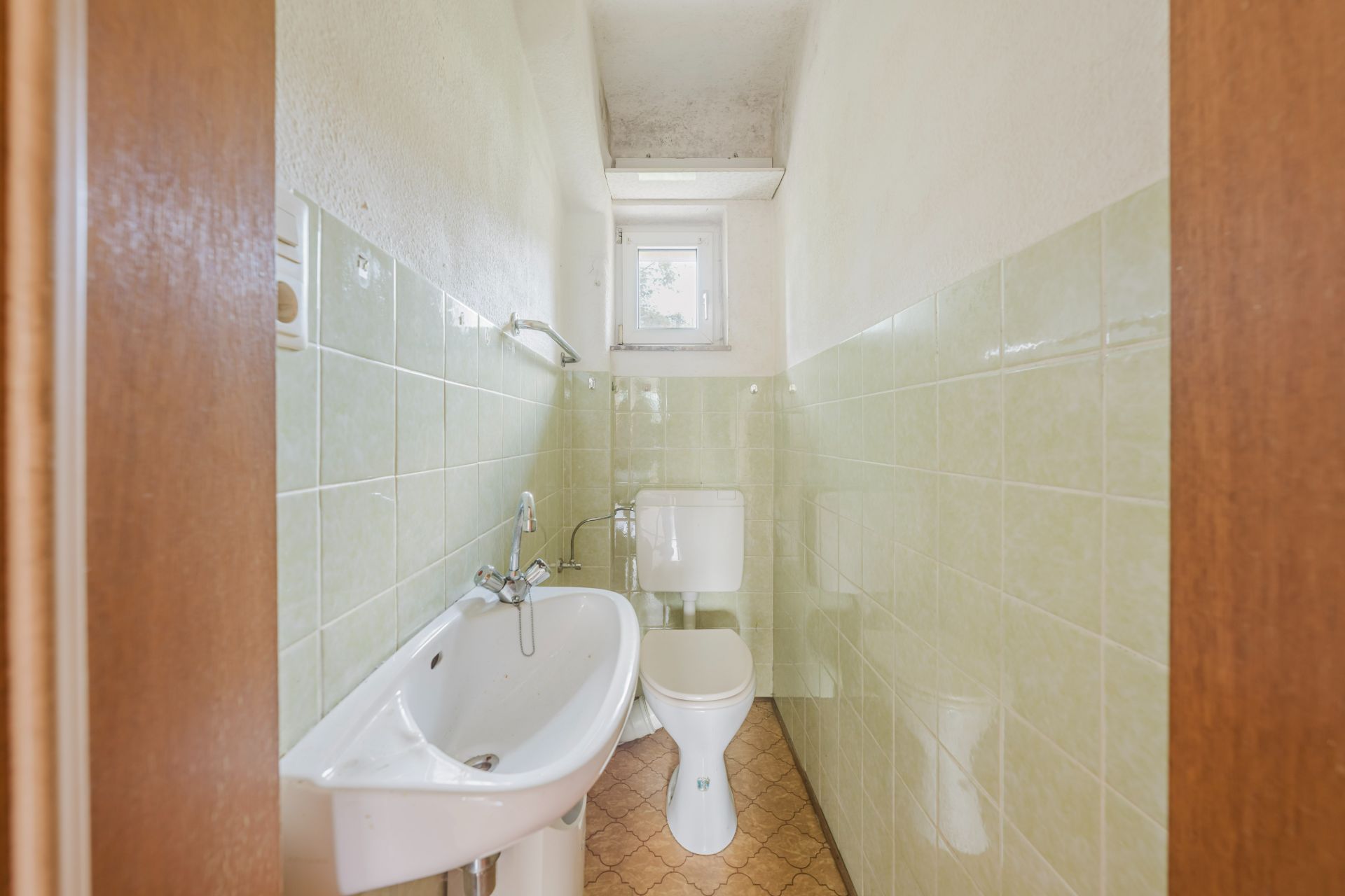 Schmales Badezimmer mit Waschbecken, Toilette, kleinem Fenster und grünen Wandfliesen.