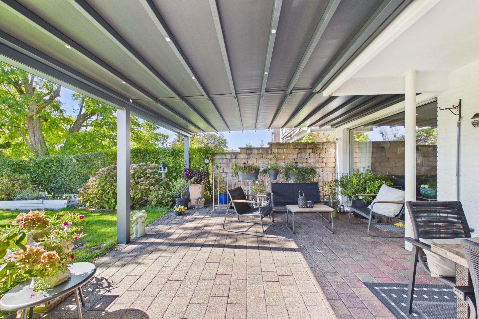 Überdachte Terrasse mit Sitzmöbeln, Pflanzen und Blick auf einen Garten.