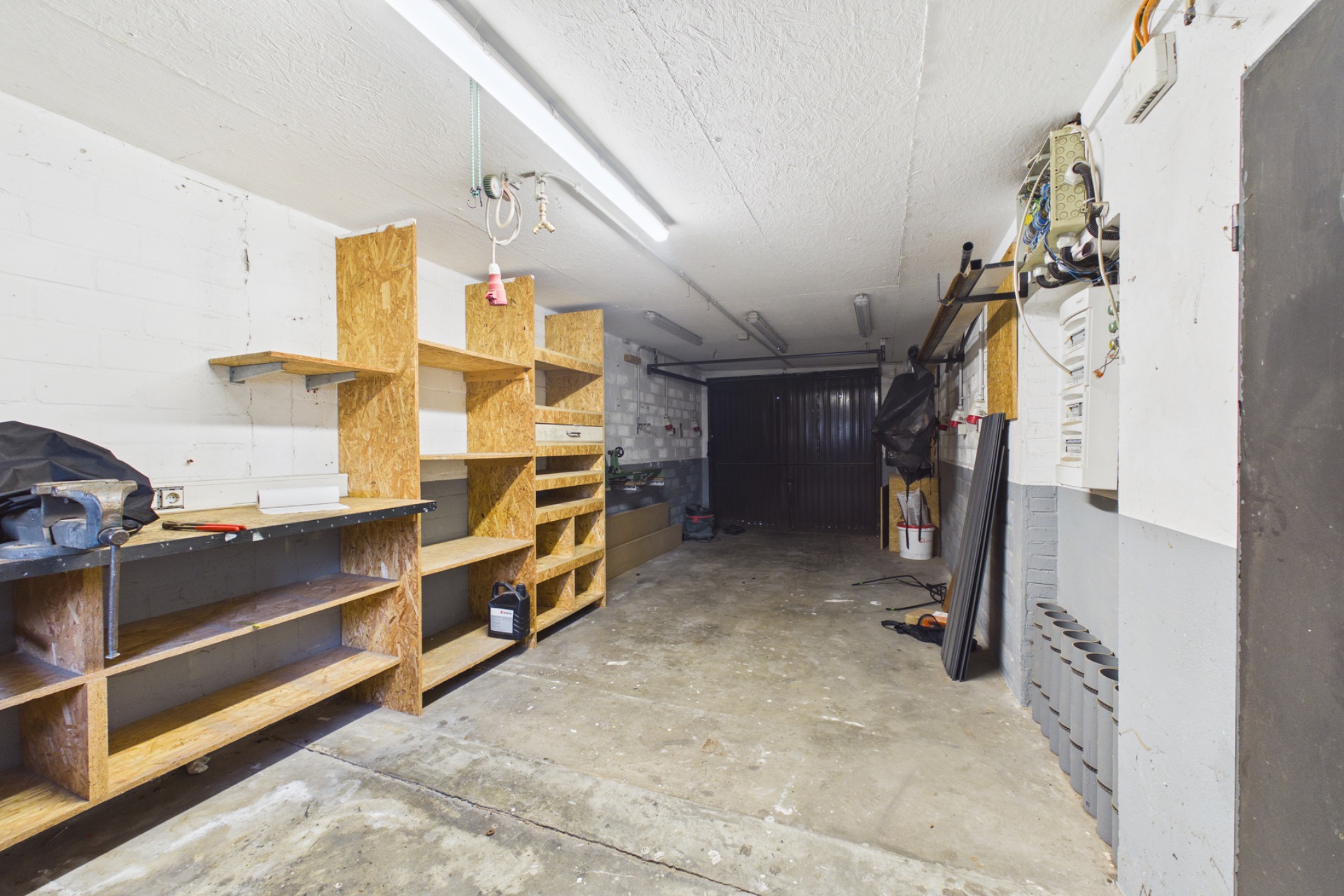 Garage mit Holzregalen, Werkzeugbank und Betonboden, an der Wand hängen Werkzeuge.