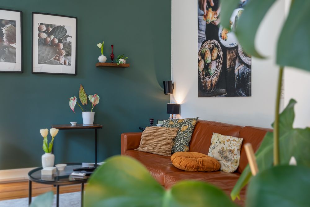 Wohnzimmer mit braunem Sofa, Pflanzen, grüner Wand und Bildern an der Wand.