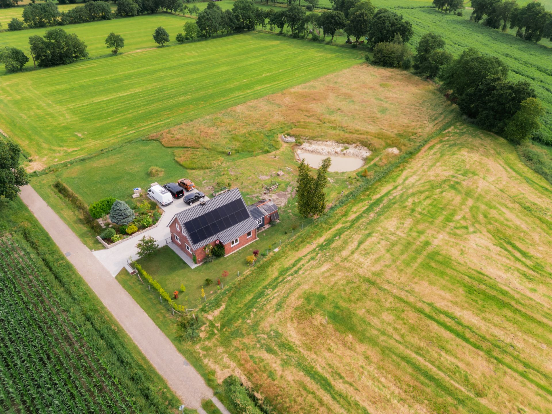 Luftaufnahme eines Einfamilienhauses mit Solardach, Garten, Teich und umgebenen Feldern.