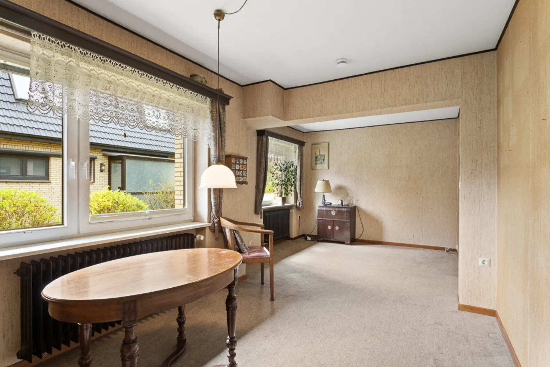 Wohnzimmer mit großem Fenster, Holztisch, Stuhl, Sideboard und Stehlampe.