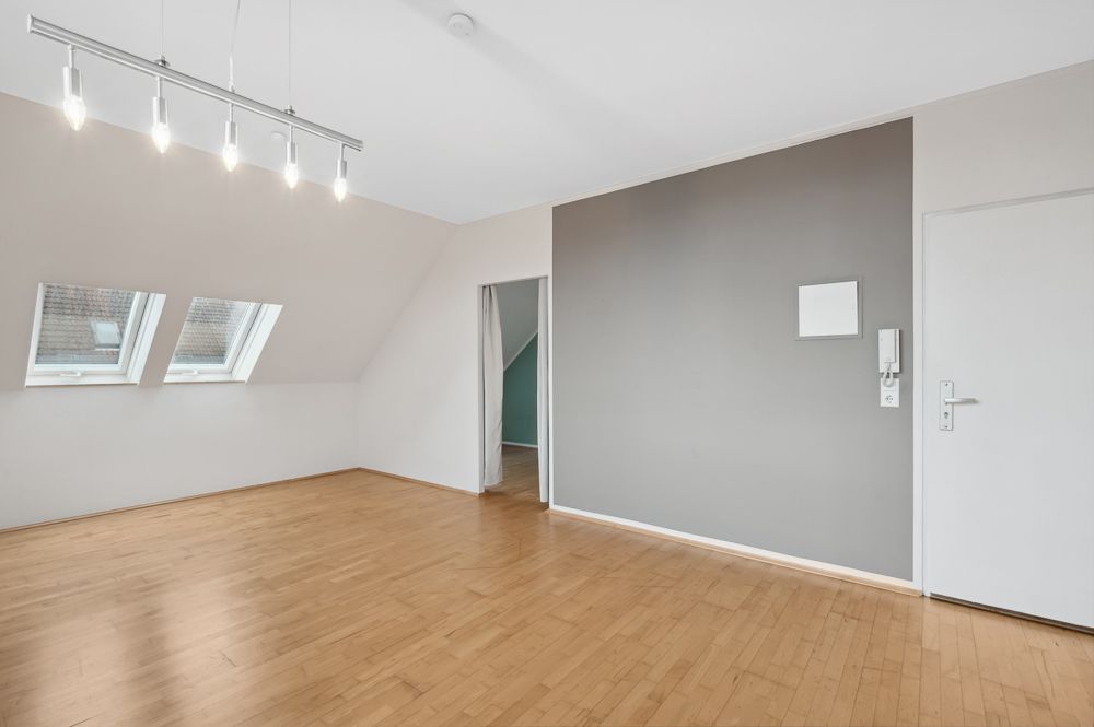Leerer Raum mit Holzfußboden, zwei Dachfenstern und grauer Wand mit Tür.