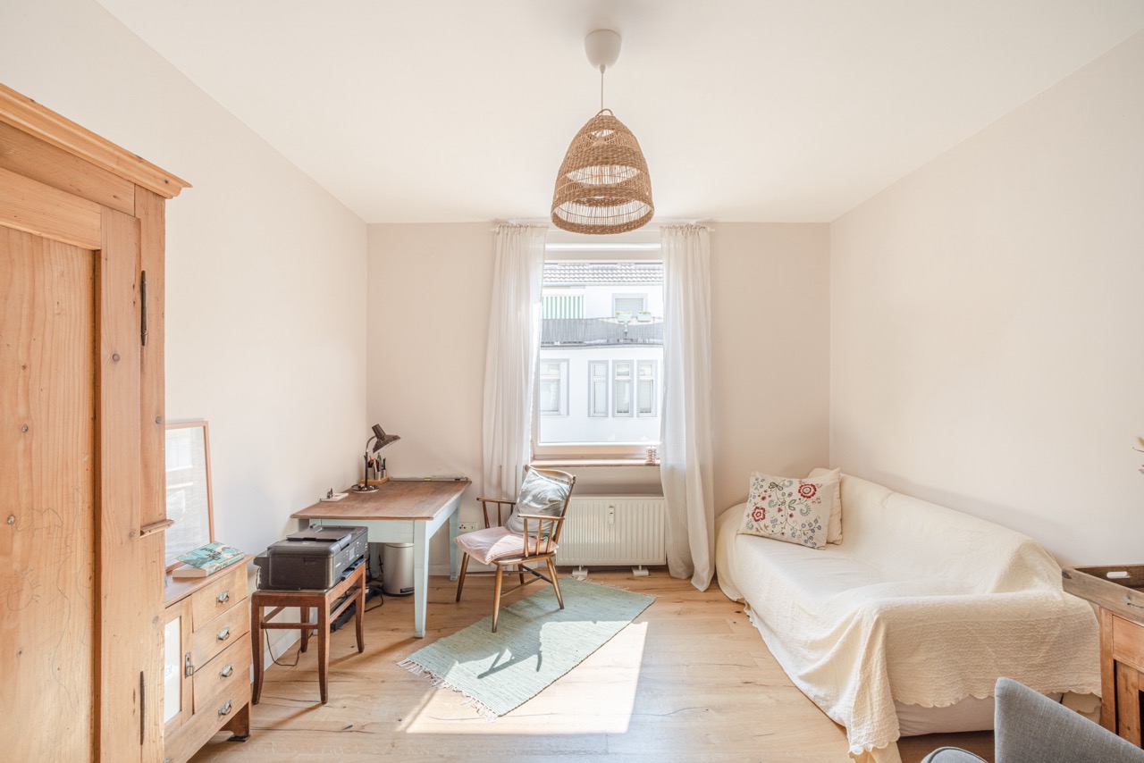 Heller Raum mit Sofa, Schreibtisch, Sessel und Holzschrank, großes Fenster mit weißen Vorhängen.