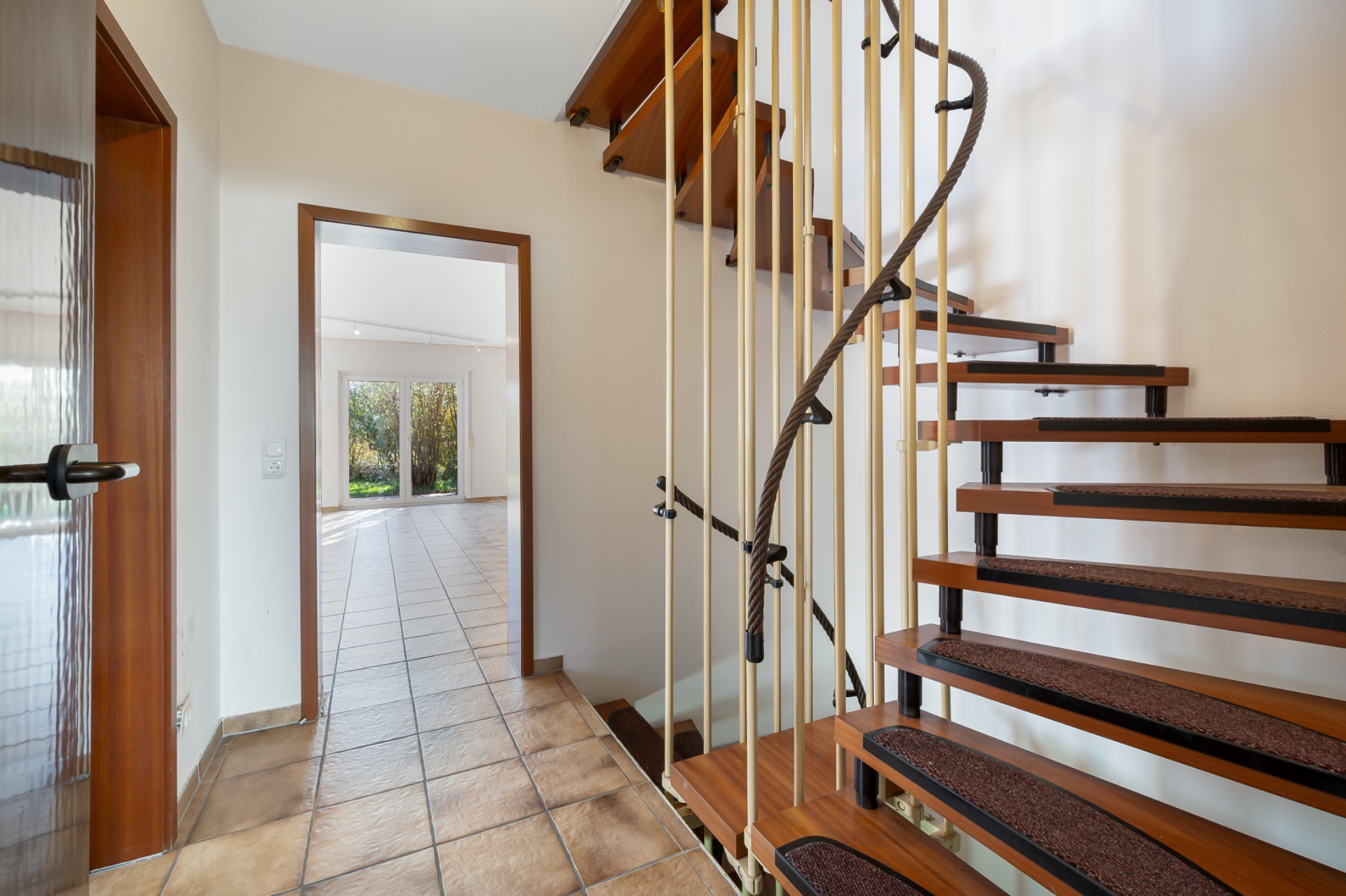 Flur mit offener Holztreppe und Blick in ein helles Wohnzimmer mit Fliesenboden.