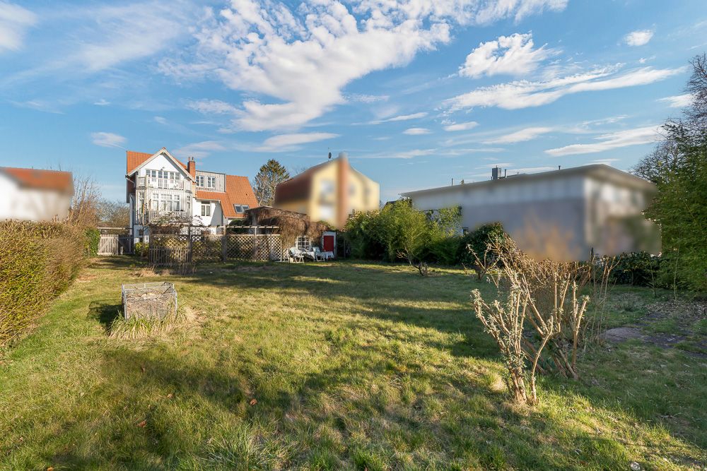 Großer Garten mit Rasenfläche, Sträuchern und einem Haus im Hintergrund bei blauem Himmel.