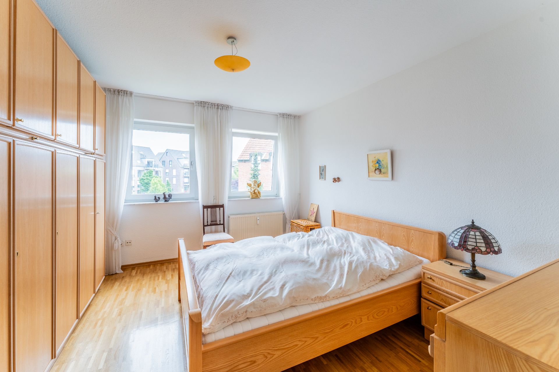 Helles Schlafzimmer mit Holzbett, Kleiderschrank, Nachttisch und zwei Fenstern.