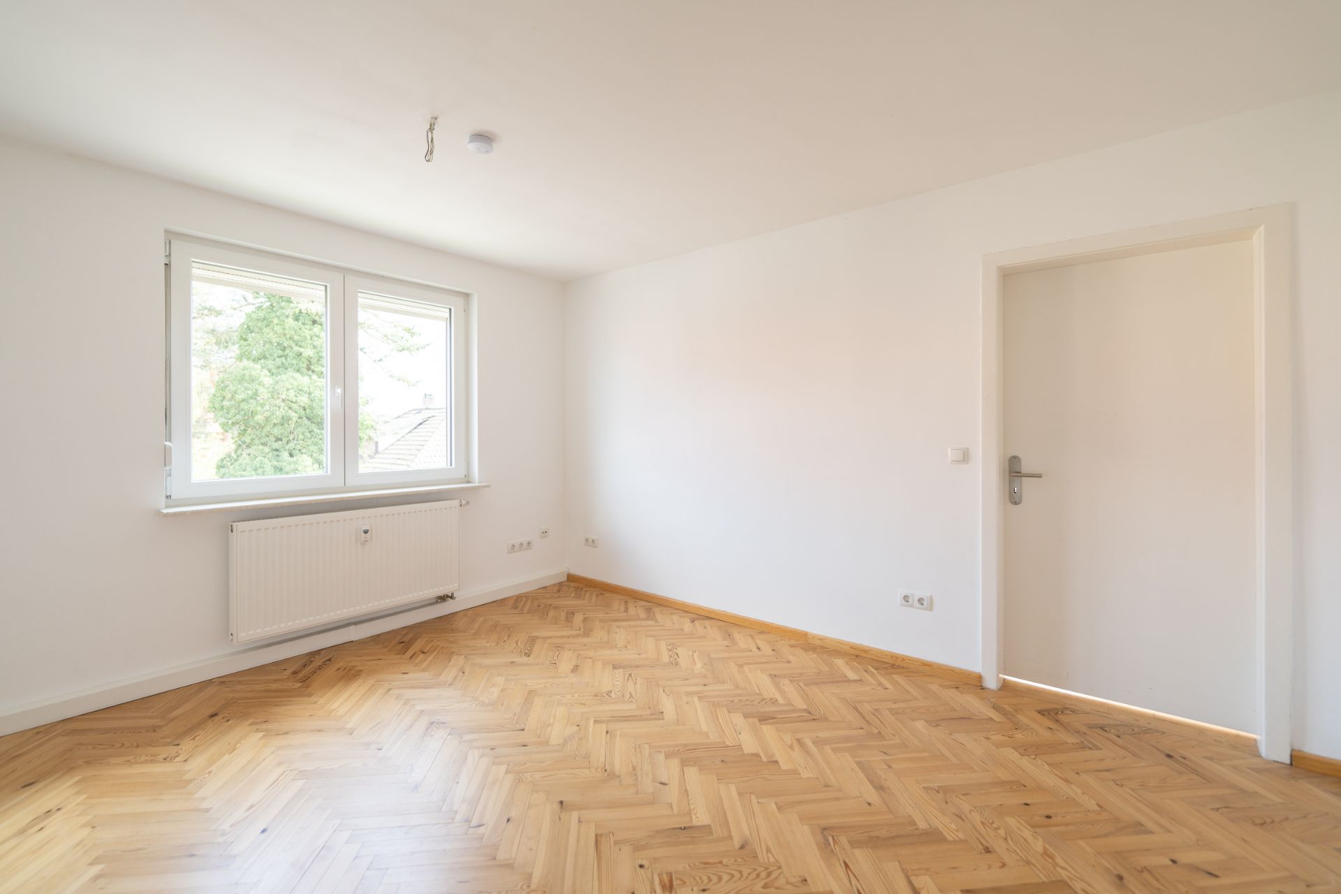 Helles, leeres Zimmer mit Parkettboden, weißer Wand, Fenster und Tür.