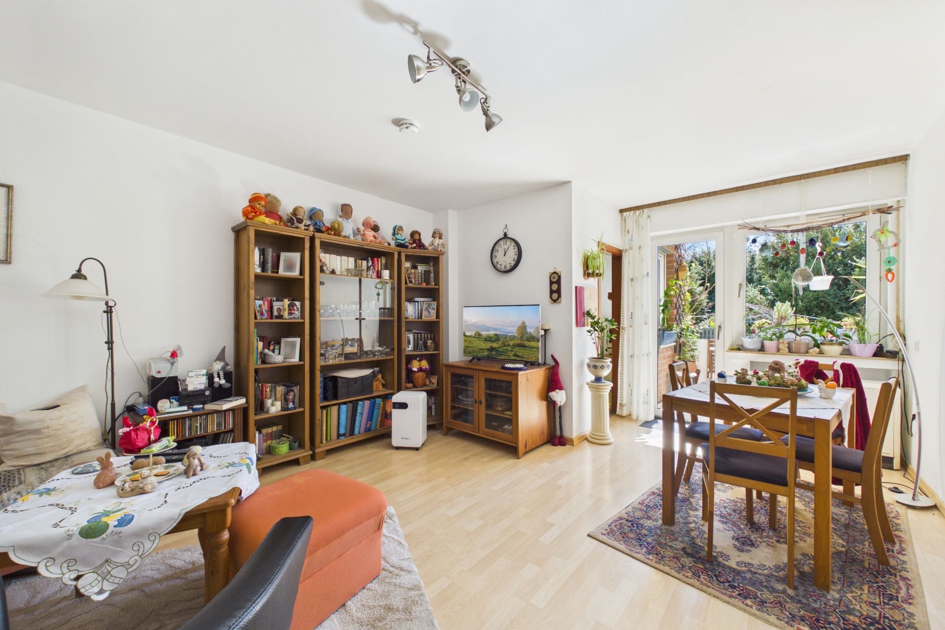 Helles Wohnzimmer mit Esstisch, Bücherregal, Fernseher und Zugang zu einem Balkon mit Pflanzen.