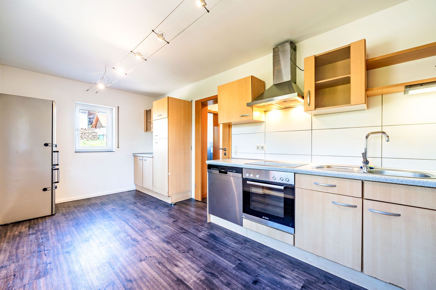 Moderne Einbauküche mit Holzfronten, Edelstahlgeräten und Fenster, auf dunklem Boden.