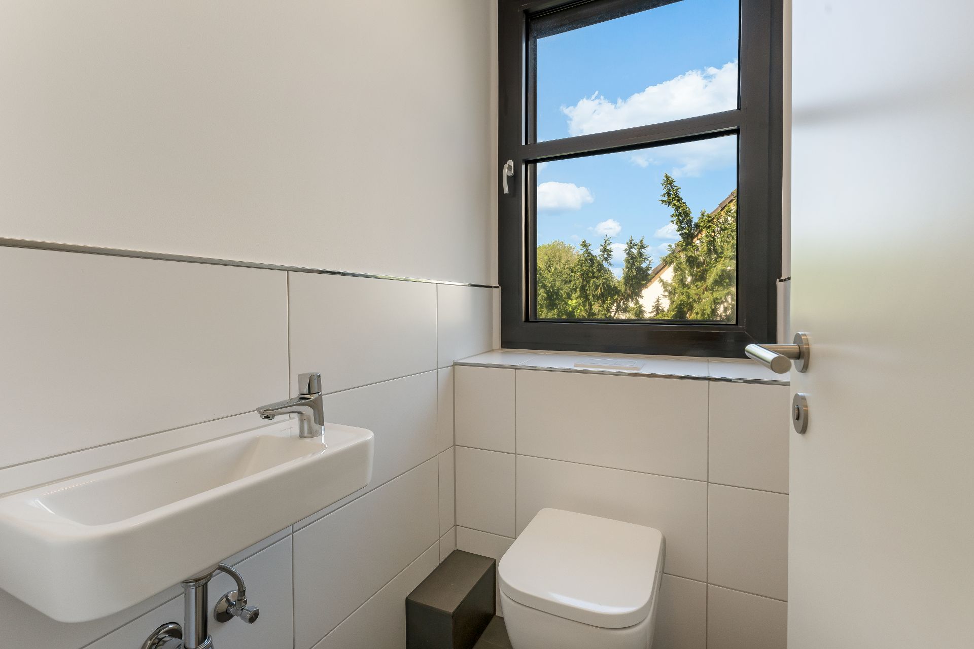 Modernes, helles WC mit kleinem Waschbecken und Fenster mit Blick ins Grüne.