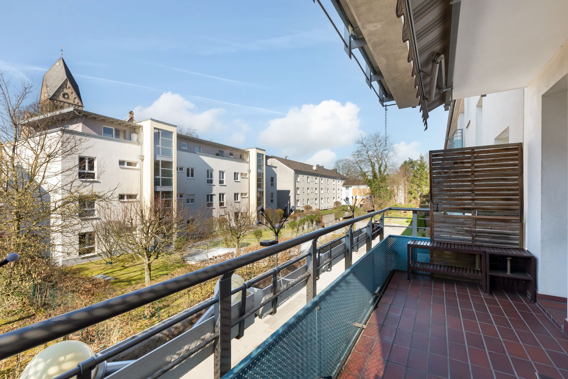 Balkon mit Blick auf Wohngebäude und Bäume, Sitzbank und Gittergeländer sichtbar.