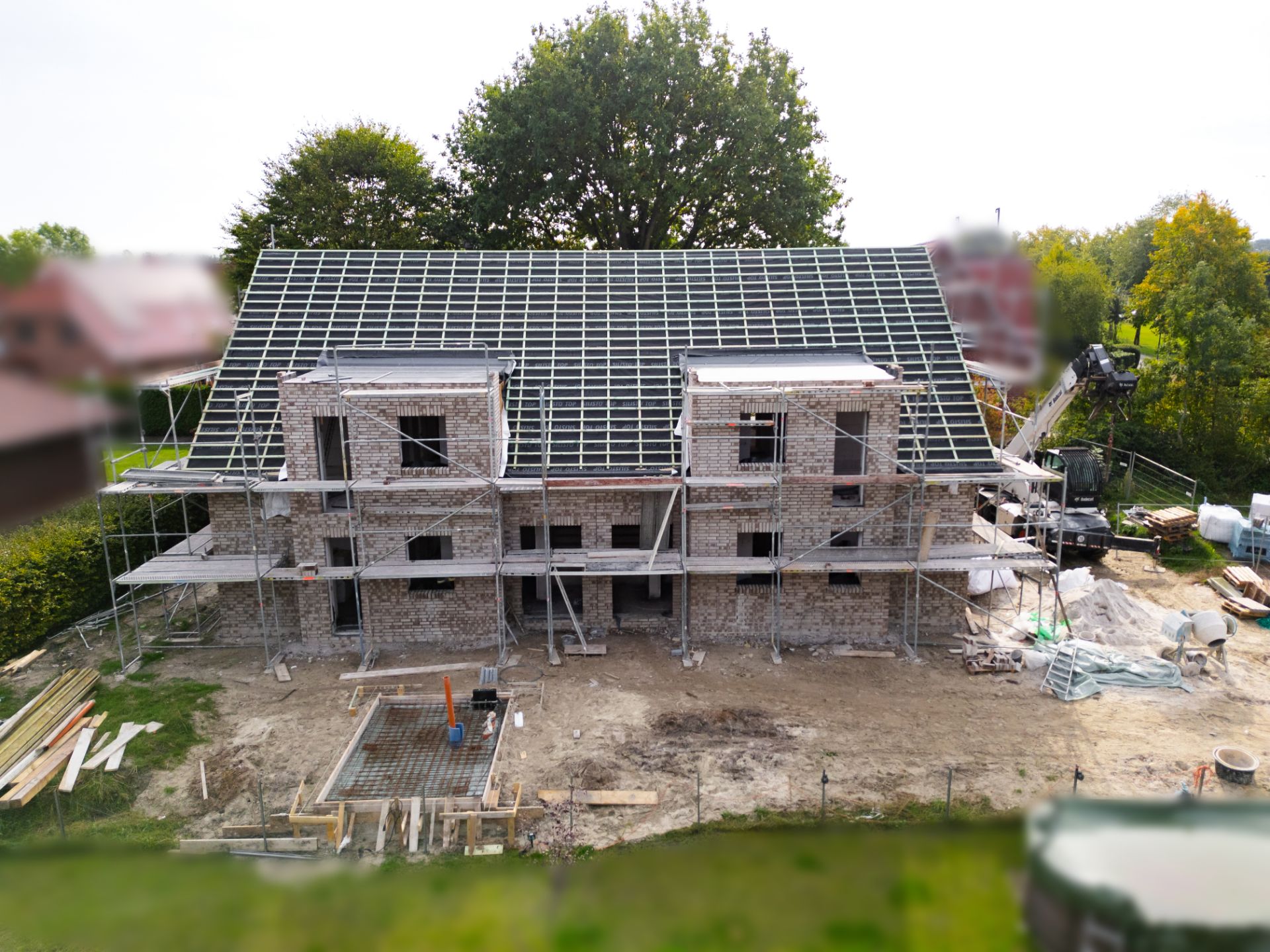 Zweistöckiges Wohnhaus im Rohbau mit Baugerüst und offenem Dach, umgeben von Baumaterialien.