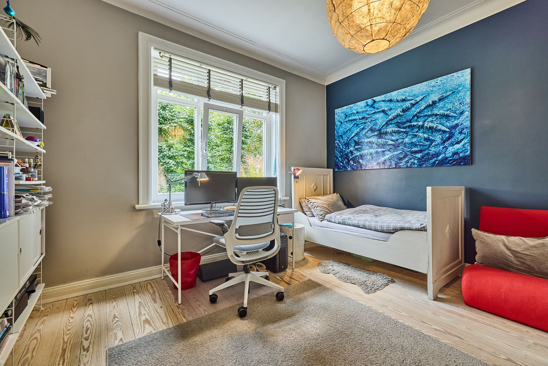 Helles Zimmer mit Schreibtisch, Stuhl, Bett, Regal und großem Fenster im Hintergrund.