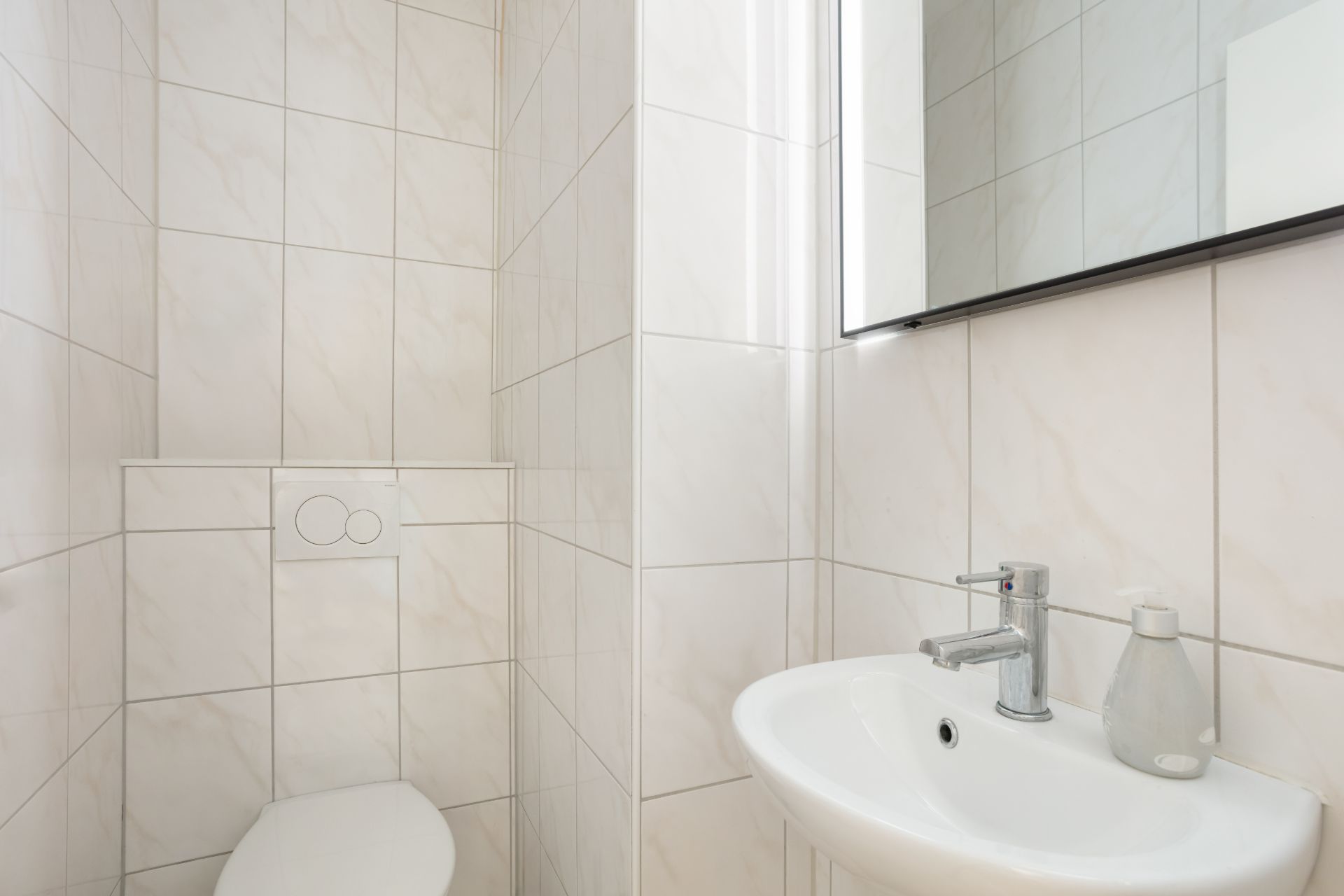 Kleines, weiß gefliestes Bad mit Wandtoilette, Waschbecken und Spiegel.
